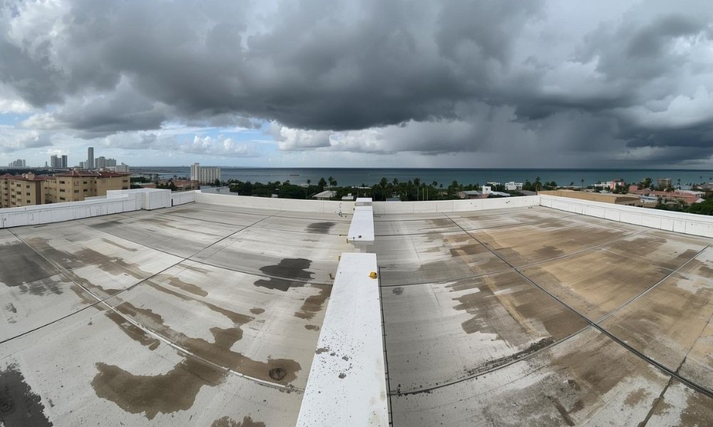 A panoramic view of a large commercial flat roof in South Florida showing the complex realities of weather stress: persistent ponding water near drains, slight discoloration from UV exposure, and heavy rain clouds moving in over a nearby coastal cityscape.