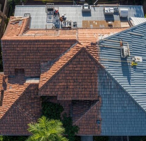 A complex South Florida residential roof replacement project in progress, captured from above. The scene displays various cost factors: a large crew working with safety gear, multiple roof faces at different pitches, and distinct sections of both concrete tile and standing-seam metal roofing materials under bright sun, framed by palm trees.