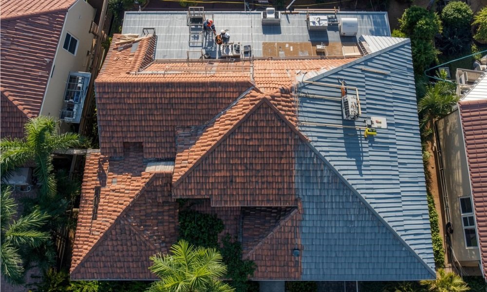 A complex South Florida residential roof replacement project in progress, captured from above. The scene displays various cost factors: a large crew working with safety gear, multiple roof faces at different pitches, and distinct sections of both concrete tile and standing-seam metal roofing materials under bright sun, framed by palm trees.