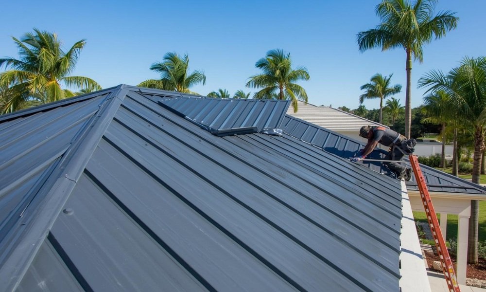 A professional photograph of an attractive, modern charcoal grey standing seam metal roof being installed on a new Florida home, demonstrating the sleek aesthetic and robust construction. A roofer is securing a panel, with palm trees and a clear blue sky in the background.